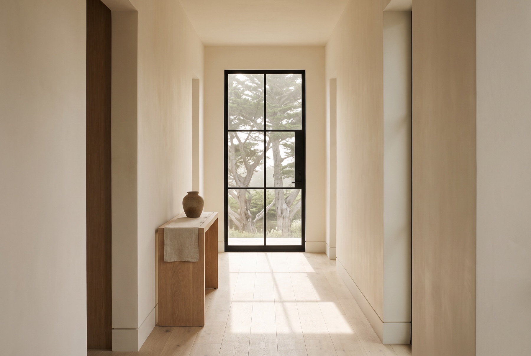 An entry hall with white oak floors, warm plaster walls and a tall steel window casting a long rectangle of light across the floor.