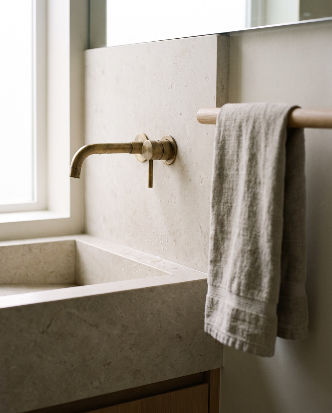 A close detail of an unlacquered brass wall-mounted tap over an integrated limestone basin.