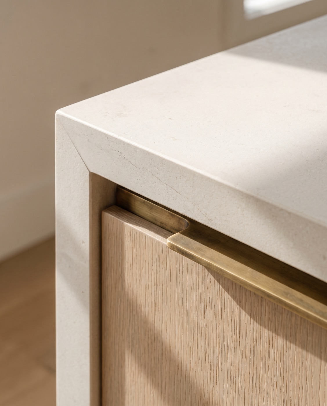 A close detail of a mitred limestone countertop edge meeting white oak cabinetry with a slim brass pull.